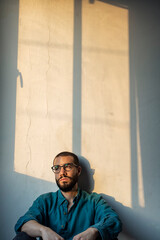 Handsome young bearded man with vintage glasses sitting against a croocked wall with a melancholic emotion in his face as the evening sun kiss him