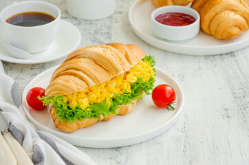 Croissant with scrambled eggs, lettuce and cherry tomatoes on a white plate with a cup of coffee on a wooden background. Horizontal, copy space.