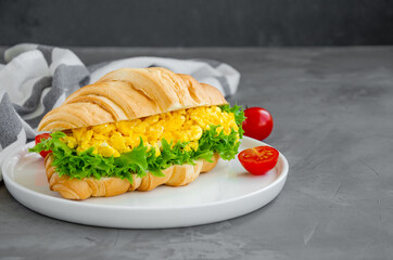 Croissant with scrambled eggs, lettuce and cherry tomatoes on a white plate with a cup of coffee on a dark concrete background. Horizontal, copy space.
