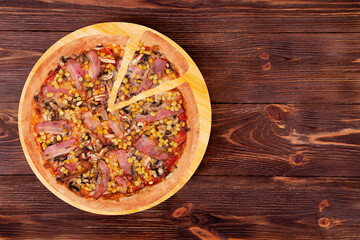 Pizza with chicken breast, corn, bacon and mushrooms, with a slice slightly removed on a wood plate which is on wooden rustic table, top view and copy space
