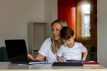 Fototapeta na wymiar Young Caucasian single mother working on laptop while her son is using digital tablet.Mother and her son spending time together at home.Mother using laptop and her son using tablet.Technology concept.