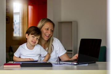 Young mother and son sitting at table and using laptop at home.Smiling mom working at home with her child while writing an email.Young woman teaching little boy to use the computer.Technology concept.