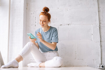 Fototapeta na wymiar Happy redhead young woman wearing wireless earphones listening relaxed pleasant music using cell phone. Cheerful smiling girl sitting near big panoramic window and looking away, on white background.