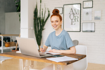 Smiling happy redhead young woman student is writing in documents, working on laptop while sitting at the desk in light cozy living room at home office, looking at camera. Concept of remote working.