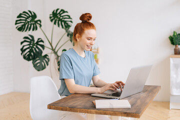 Cheerful smiling redhead young business woman is working on laptop computer at the desk in light cozy room at the home office.