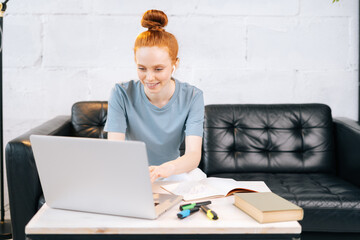 Smiling redhead young business woman is working on laptop at home office. Happy female wearing earphones listening music in cozy light room. Cute girl typing on notebook in self-isolation.