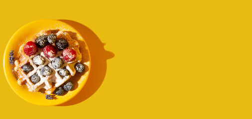 Belgian waffel with raspberries, blueberries, blackberries seasoned with powdered sugaron yellow saucer. Yellow background. Flatly. Hard light. Copy space.