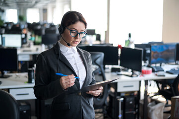 Serious business woman in a suit answers the customer's call on the headset. Female manager takes notes while talking with a client. Administrator at work in the office.