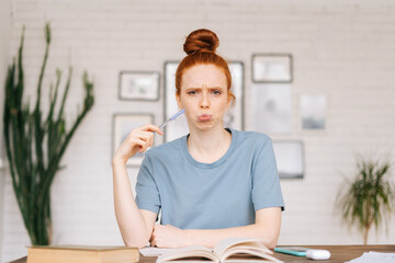 Dissatisfied bored redhead young woman student sitting at a table with textbooks and a workbook, looking at camera. Girl student preparing for college exams.