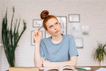 Pensive redhead young woman student is preparing for exam read textbook noting in notebook at desk at home office. Girl doing her homework at home.