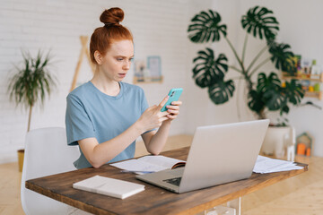 Serious redhead young businesswoman is using cell phone while sitting at the desk in light cozy living room at home office.