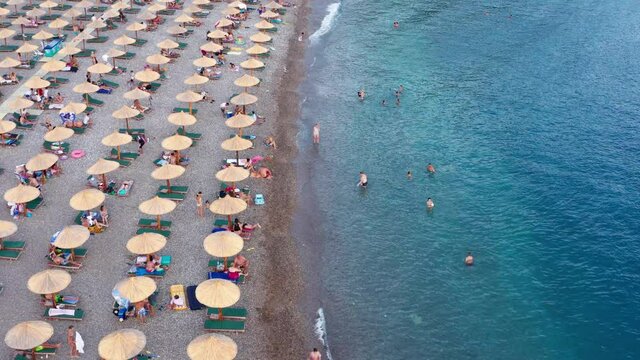 Rows of reed umbrellas at the sandy beach in Canj, on the Adriatic coast of Montenegro. People on vacation under sunshades, or in blue and turquoise sea. Impact of coronavirus pandemic on tourism.
