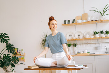Smiling redhead young woman sitting in lotus pose at the desk and looking away. Happy girl wearing white casual clothes meditating at home office. Concept of rest during remote work in self isolation.