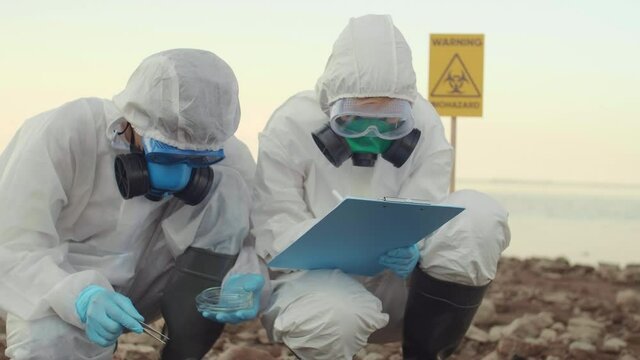 Medium shot of male and female ecology experts wearing protective suits sitting on ocean shore, taking sample of its soil using Petri dish and tweezers and writing down results in document