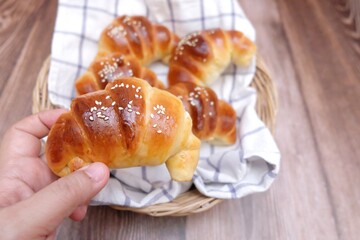 A woman hand holding a croissant with blurred a group of homemade croissants in a basket and old wooden dinning table background