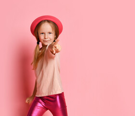 little girl in stylish blouse, leggings and panama pointing finger at camera. Cropped shot isolated on pink, copy space