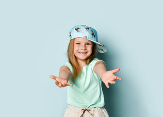 Adorable little girl wearing cap with pineapple pattern extending her arms with smile going to embrace a spectator. Studio shot isolated on light blue