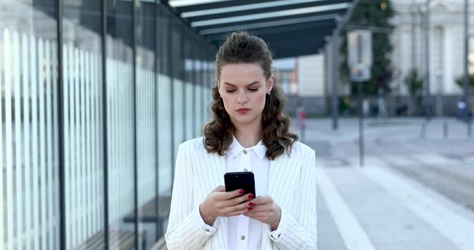 Front view of concentrated young woman in trendy business suit working on smartphone outdoors. Concept of busyness, successful career and technology.