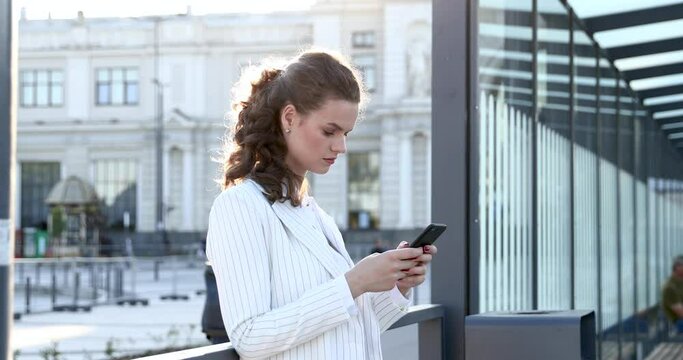 Beautiful young woman in trendy white suit standing outdoors and using modern smartphone for chatting with business partners. Concept of people and technology.