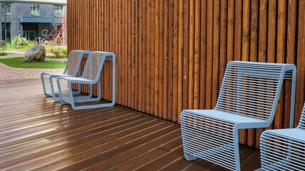 Resting point inside residential area courtyard in rainy day. Two wet metal chairs with raindrops of modern design close-up on a background of wet wooden shield.Comfortable urban public furniture.