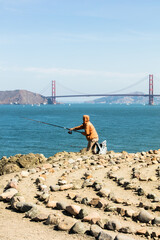 Fisherman casting line into San Francisco Bay with Golden Gate Bridge in background. Foreground is the secret place of rocks, the labyrinth zen-like maze.
