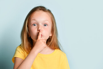Fototapeta premium Blonde child in yellow t-shirt. She is showing be quiet sign by her forefinger while posing against turquoise studio background. Close up