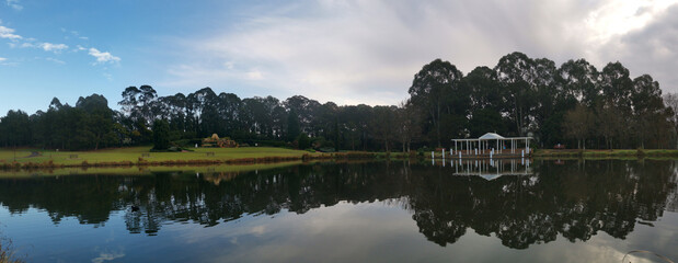 Obraz premium Beautiful morning panoramic view of a still pond in a park with stunning reflections of blue puffy sky and tall trees, Fagan park, Galston, Sydney, New South Wales, Australia