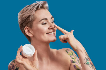 Portrait of beautiful half naked tattooed woman with short hair smiling, while applying cosmetic...