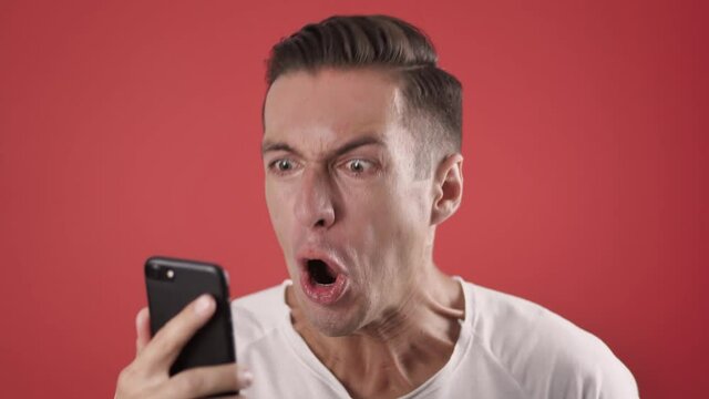 Angry Man Using Smartphone on red background. Man is angry with the message that he read in the smartphone. Angry man uses smartphone.