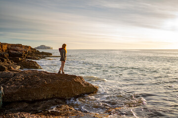 Side view of woman looking at seascape while standing on rock against sky during sunset