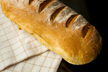 Freshly baked white bread with selective focus on one side rests on a glossy black surface and a checkered light towel.