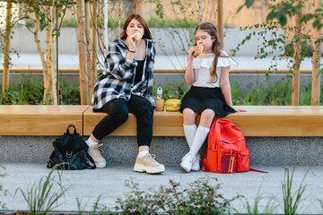 Two schoolgirls, a child and a teenager, have their lunch on a bench in the schoolyard.