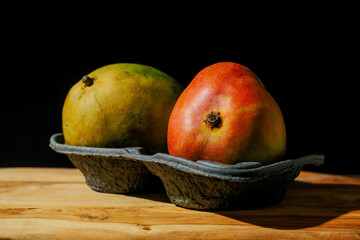 Two fresh whole mango fruit in a black plastic container on a wooden surface, Black background, Fresh fruit retail concept.