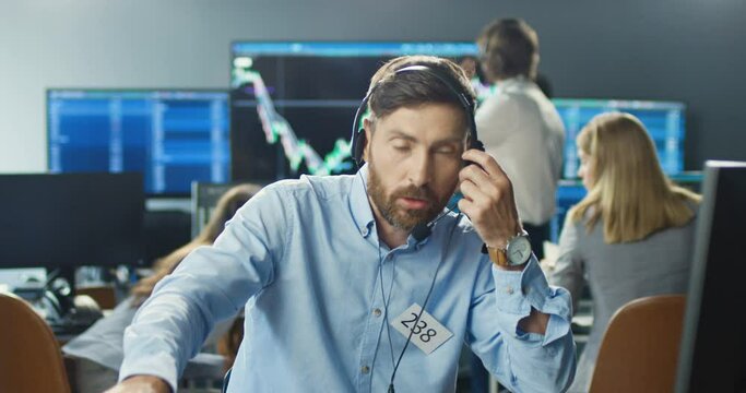 Caucasian handsome male broker in headset working at computer in trading office and answering phone calls on many telephones. Trader buying and selling while talking with clients and working hard.