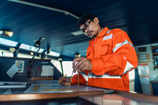 Filipino deck Officer on bridge of vessel or ship wearing coverall during navigaton watch at sea . He is plotting position on chart