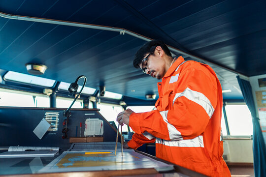 Filipino deck Officer on bridge of vessel or ship wearing coverall during navigaton watch at sea . He is plotting position on chart