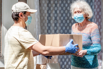 Young male volunteer in mask gives an elderly woman boxes with food near her house. Son man helps a...