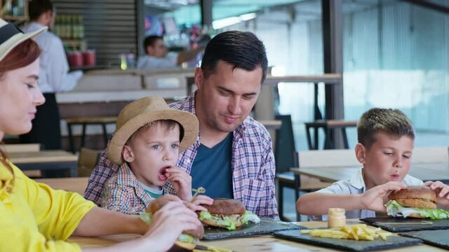 happy cheerful man and woman with male children enjoy eating tasty juicy burgers with fries sitting at table in cafe