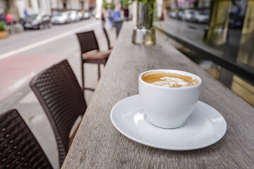 Selected focus and close up view of a white ceramic cup of cappuccino on top with beautiful delicate latte art with leaf pattern, on wooden outdoor rough counter of coffee shop on the sidewalk.
