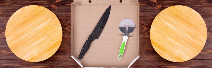 Two empty wooden pizza plates and a open pizza box with kitchen utensils in it lie on the wooden table. Flat lay, pizza cutter and a knife, top view