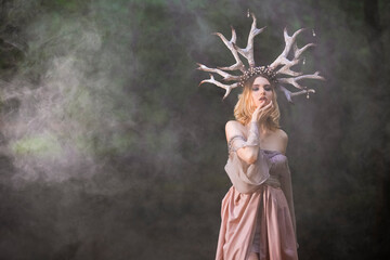Young Beautiful Girl Posing With Artistic Deer Horns  In Summer Forest and Wearing Light Dress as  Forest Nymph Concept. Colorful Artistic Smoke Used. Art Photography.