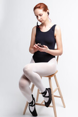 Portrait of Female Ballet Dancer in Body Suit Sitting On Chair and Listening Music on Headphones. Against White Background.