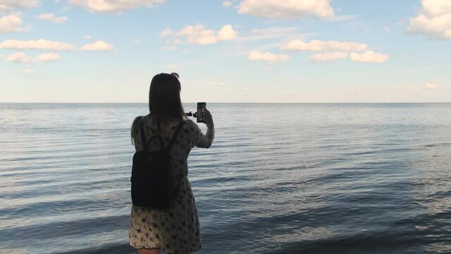 Back view portrait of woman making selfie in the background of beautiful seascape using a smartphone and the monopod at sunset