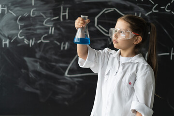 little girl student with glasses in a laboratory coat stands near the chalkboard. Holds a chemical...