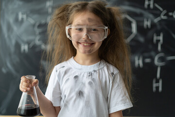 Cute little girl near the blackboard at a chemistry lesson in class. Hold the flask with the blue liquid in your hands. happy face