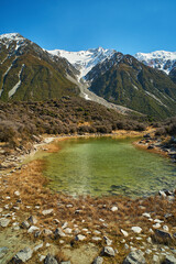 Scenic view of overlooking a beautiful tarn lake surrounded my rocks and alpine grass with snow covered mountain peaks in the background in Mt Cook New Zealand.