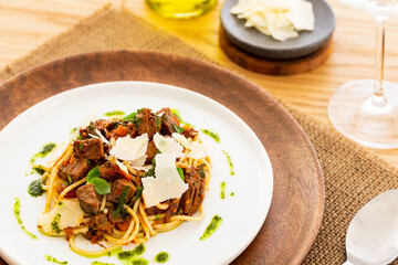 Beef ragout sauce spaghetti pasta with tomatoes, basil and pesto. Aside a spoon, a bottle of olive oil, parmesan and wine glass. White plate on a wooden plate on a jute mat on an oak table.