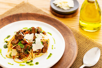 Beef ragout sauce spaghetti pasta with tomatoes, basil and pesto. Aside a spoon, a bottle of olive oil and parmesan. White plate on a wooden plate on a jute mat on an oak table.