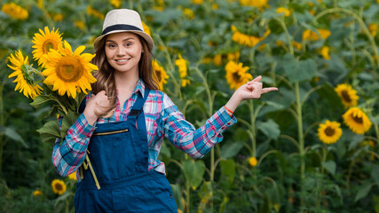 Energetic, female farmer pointing to her left while holding many sunflowers, in the middle of a green and yellow sunflower field.