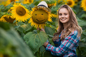 Gorgeous, young, adorable, energetic, female farmer standing near a sunflower with a smiley face, in the middle of a golden sunflower field.